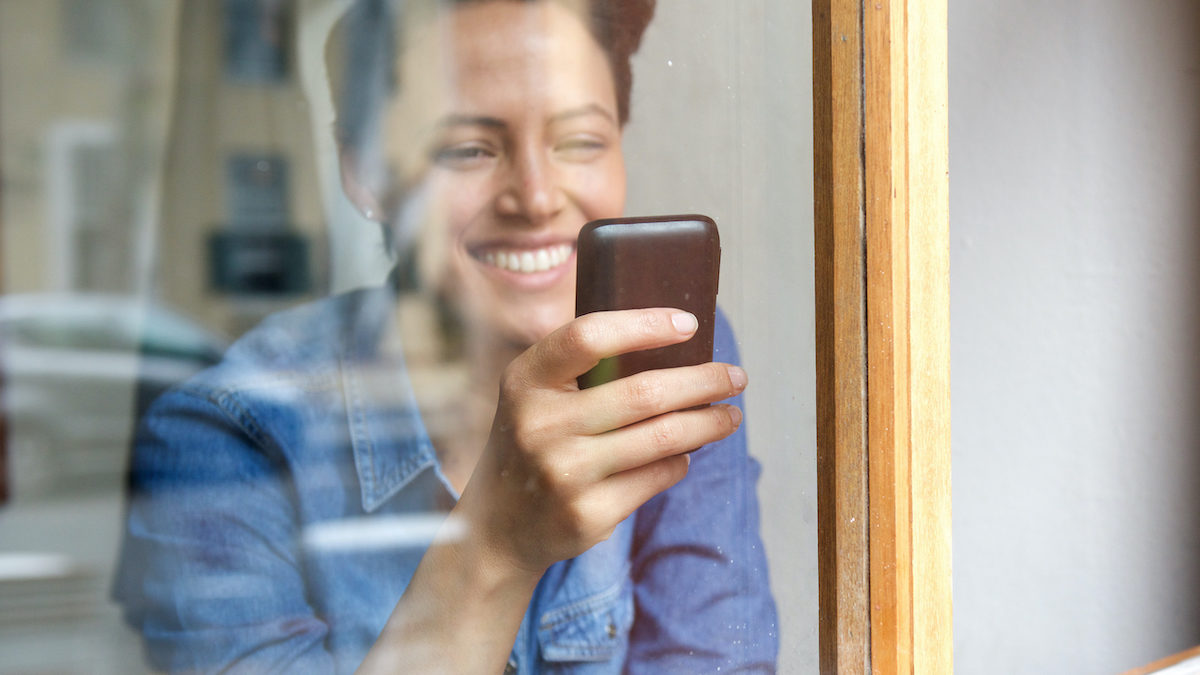 Portrait looking through window at smiling young woman using cell phone
