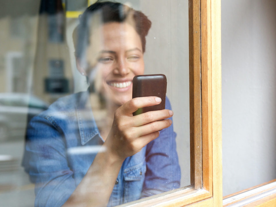 Portrait looking through window at smiling young woman using cell phone