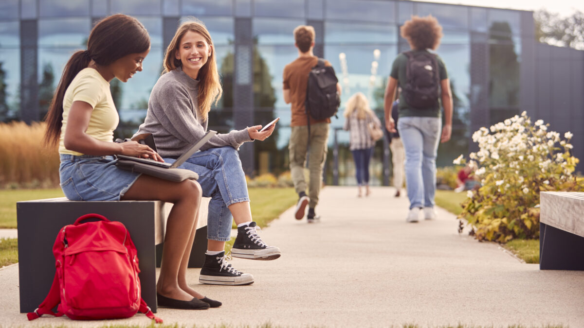 Female University Or College Students Sitting Outdoors On Campus Talking And Working On Laptop