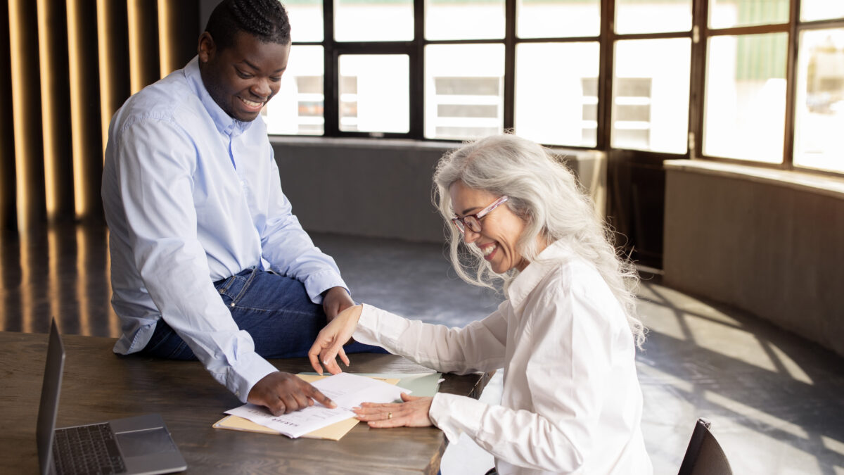 Getting New Job. Happy Black Businessman Signing Employment Contract Papers With New Employee Woman, Pointing Finger At Documents On Desk In Modern Office Indoor. Career And Paperwork