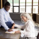 Getting New Job. Happy Black Businessman Signing Employment Contract Papers With New Employee Woman, Pointing Finger At Documents On Desk In Modern Office Indoor. Career And Paperwork