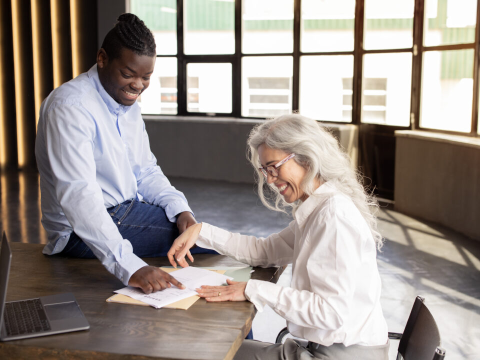 Getting New Job. Happy Black Businessman Signing Employment Contract Papers With New Employee Woman, Pointing Finger At Documents On Desk In Modern Office Indoor. Career And Paperwork