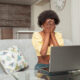 portrait African American black woman at home. Indoor, woman is frightened, shocked, unpleasantly surprised, angry. Emotions, sitting in front of laptop. covers face with hands, does not want to see