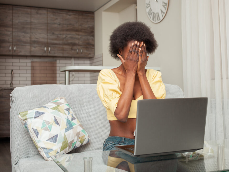 portrait African American black woman at home. Indoor, woman is frightened, shocked, unpleasantly surprised, angry. Emotions, sitting in front of laptop. covers face with hands, does not want to see