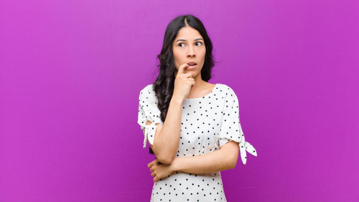 young pretty latin woman with surprised, nervous, worried or frightened look, looking to the side towards copy space against purple wall