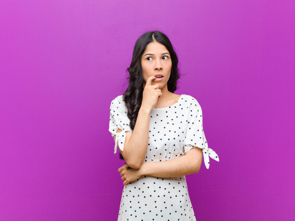 young pretty latin woman with surprised, nervous, worried or frightened look, looking to the side towards copy space against purple wall