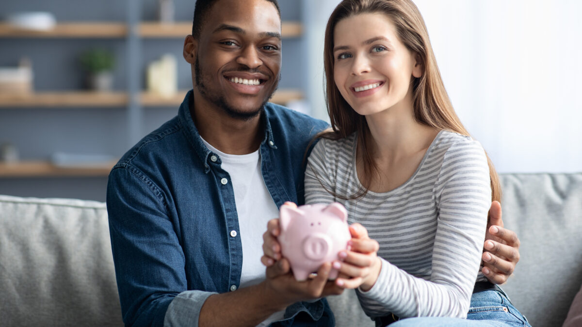 Happy Young Interracial Couple Holding Pink Piggybank In Hands And Smiling At Camera, Cheerful Multicultural Spouyses Enjoying Saving Money For Future, Sitting On Couch In Cozy Living Room