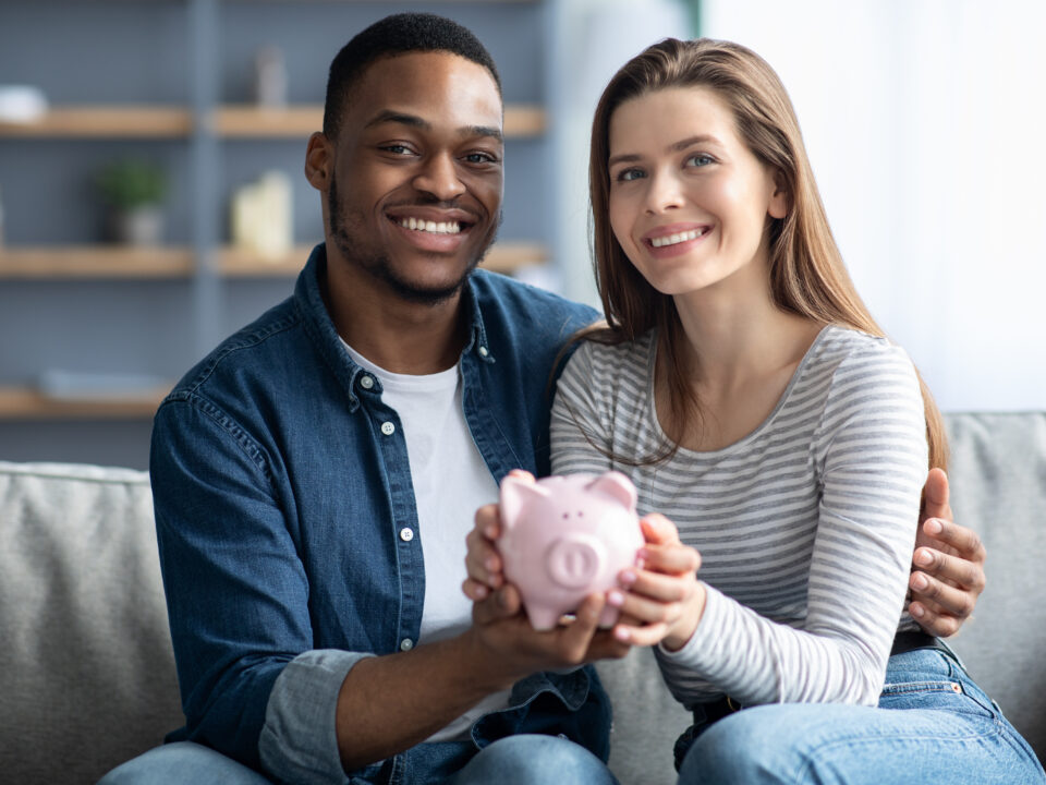 Happy Young Interracial Couple Holding Pink Piggybank In Hands And Smiling At Camera, Cheerful Multicultural Spouyses Enjoying Saving Money For Future, Sitting On Couch In Cozy Living Room