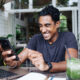 Joyful adult ethnic guy in casual clothes with watch concentrating on screen and using smartphone while sitting at laptop with notepad and glass of green drink with black straw in cafe during daytime on blurred background