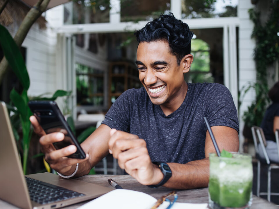 Joyful adult ethnic guy in casual clothes with watch concentrating on screen and using smartphone while sitting at laptop with notepad and glass of green drink with black straw in cafe during daytime on blurred background