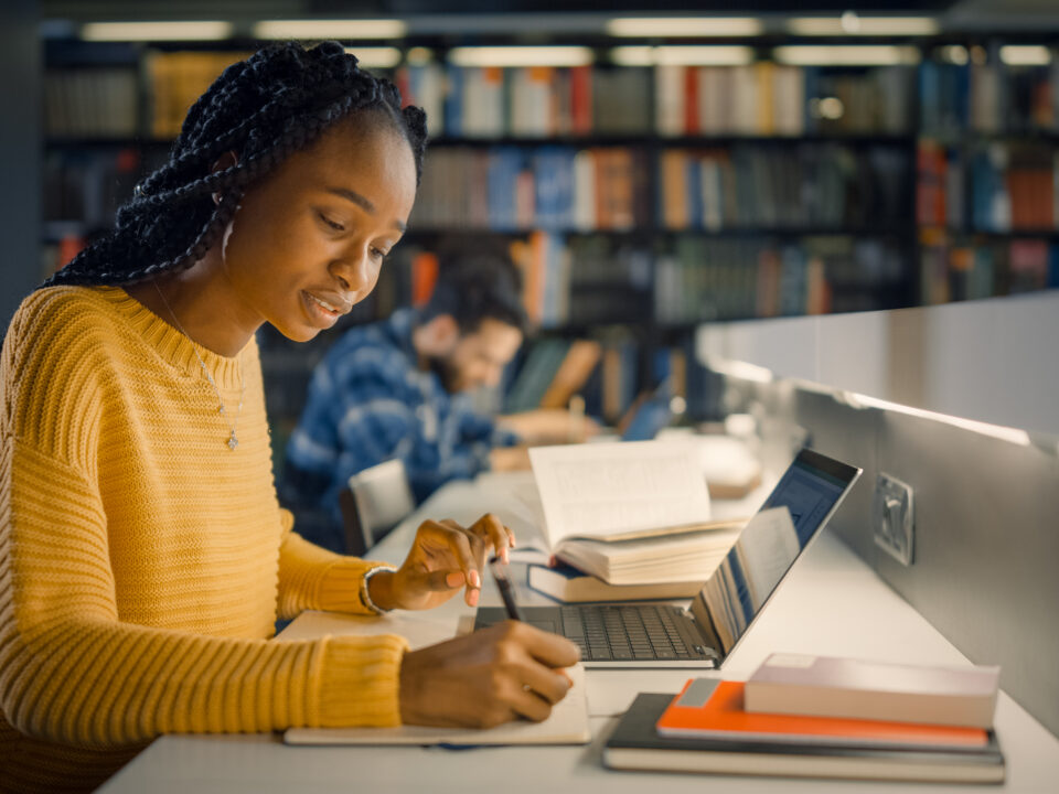 University Library: Gifted Beautiful Black Girl Sitting at the Desk, Uses Laptop, Writes Notes for the Paper, Essay, Study for Class Assignment. Diverse Group of Students Learning, Studying for Exams.