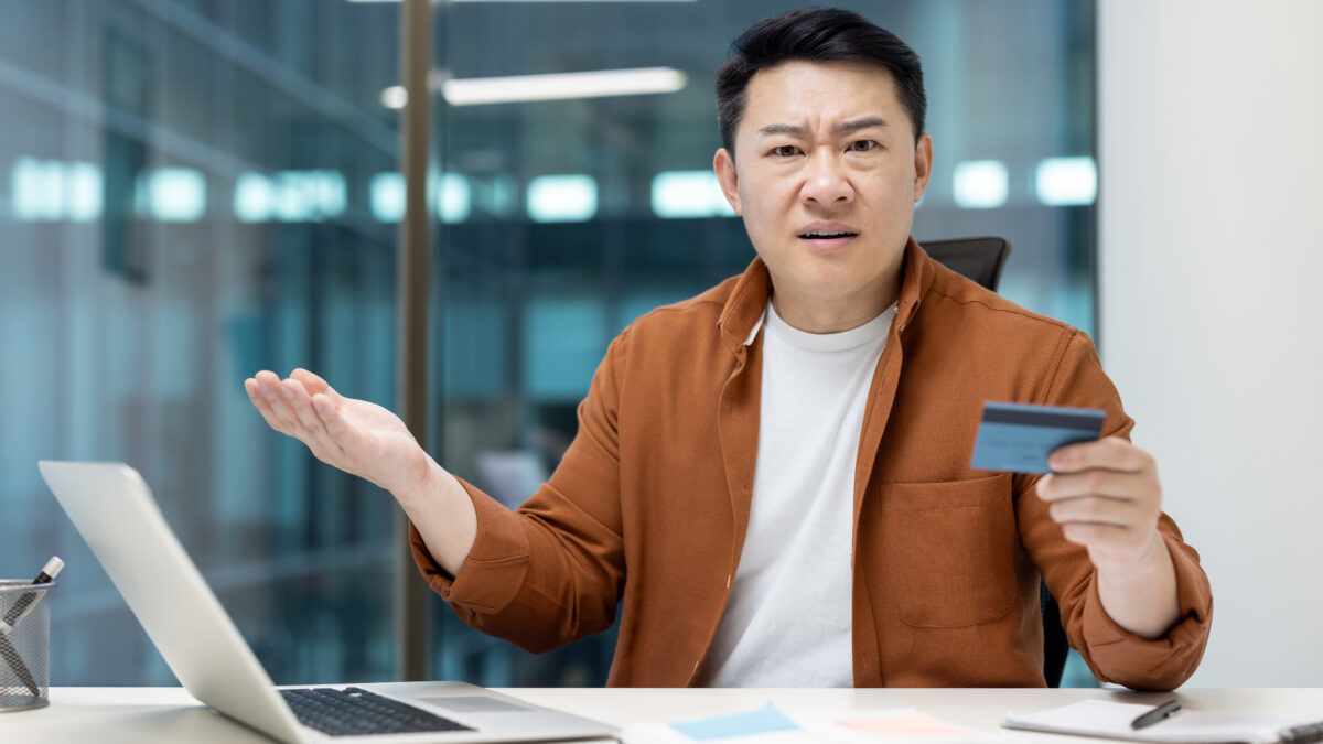 Money transfer blocking and fraud. Upset and disappointed asian man looking at camera while holding bank credit card, account access denied and error.