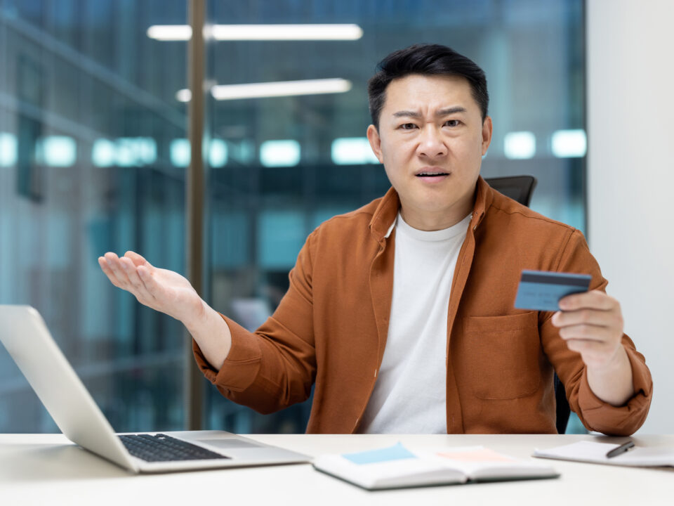 Money transfer blocking and fraud. Upset and disappointed asian man looking at camera while holding bank credit card, account access denied and error.