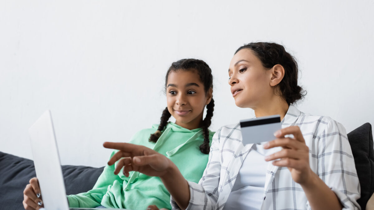 african american woman holding credit card and pointing with finger at laptop near amazed daughter