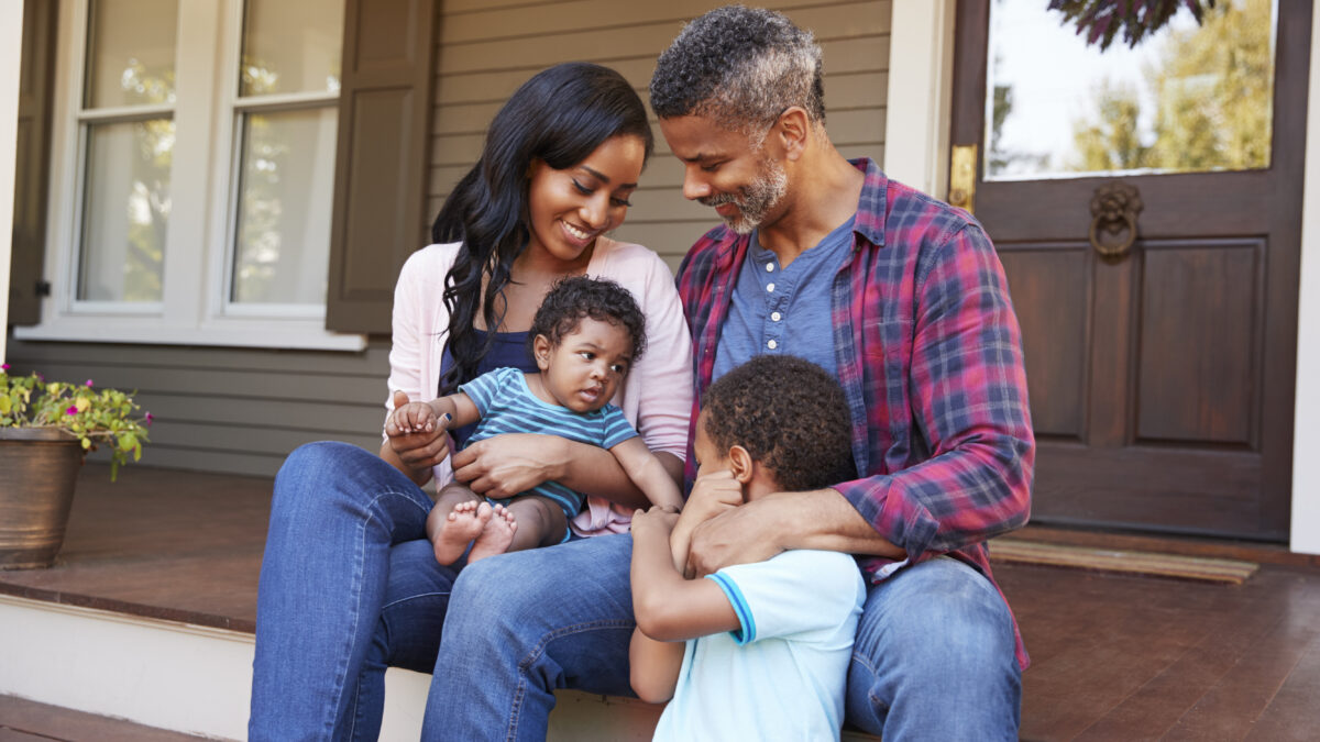 Family With Baby Son Sit On Steps Leading Up To Porch Of Home