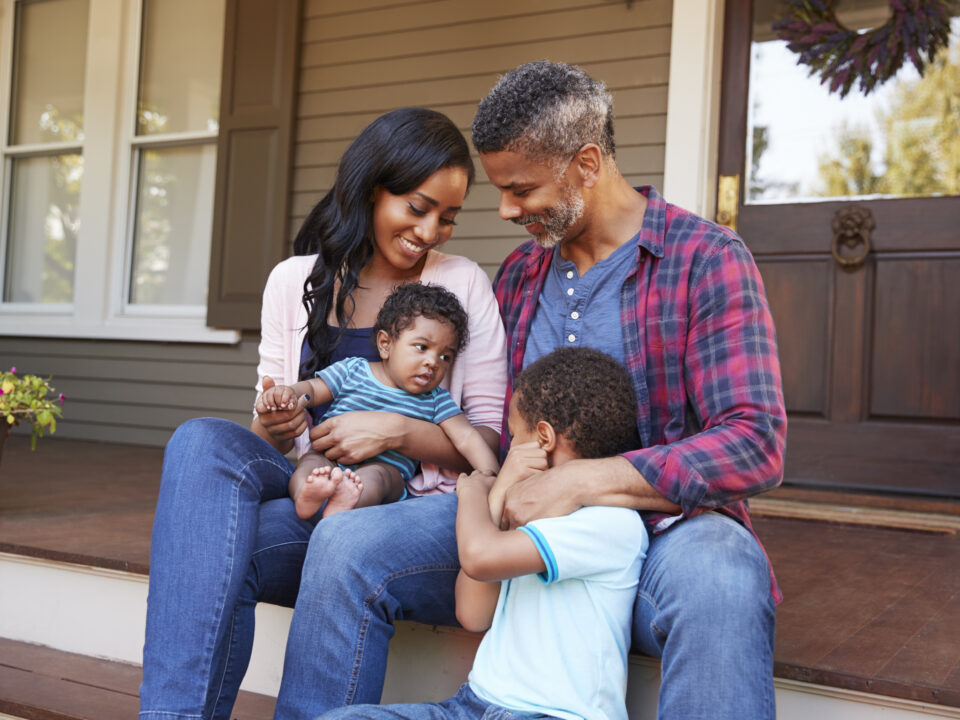 Family With Baby Son Sit On Steps Leading Up To Porch Of Home