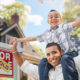 Happy Father and Son Riding Piggyback In Front of New House and Sold For Sale Real Estate Sign.