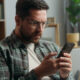 Caucasian adult man male guy at home living room nervous looking at mobile phone bad news amazed shocked close mouth with hand message from bank stressed smartphone notification online error scam