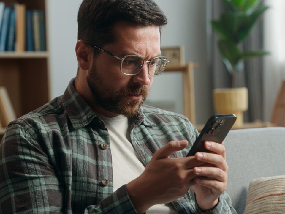 Caucasian adult man male guy at home living room nervous looking at mobile phone bad news amazed shocked close mouth with hand message from bank stressed smartphone notification online error scam