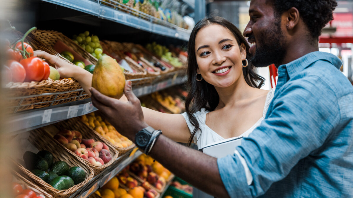 happy asian woman looking at african american man near fruits in store