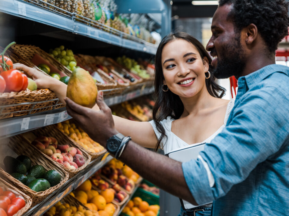 happy asian woman looking at african american man near fruits in store