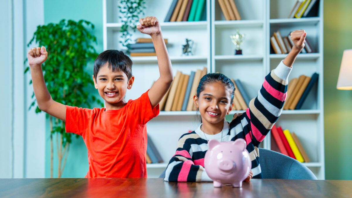 Indian siblings placing coins in a piggy bank at home, learning the concept of saving money together