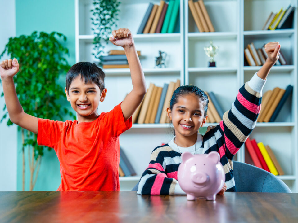 Indian siblings placing coins in a piggy bank at home, learning the concept of saving money together