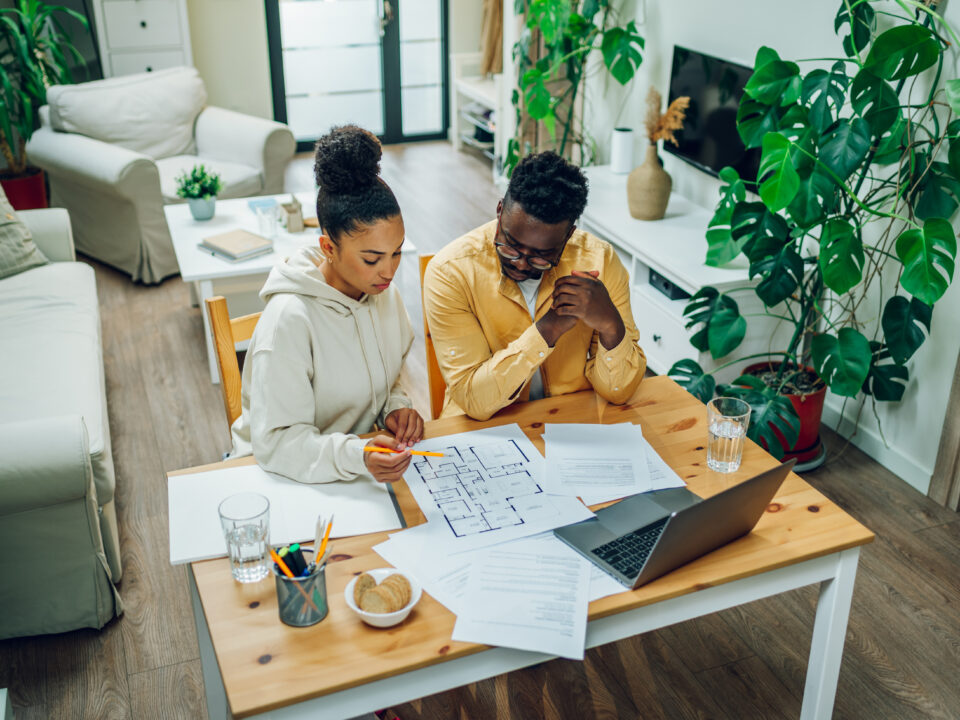 Diverse couple sitting at home office with a laptop at the table at home and looking the blueprints of their new house. Investing money into new home. Making financial strategy. Copy space.